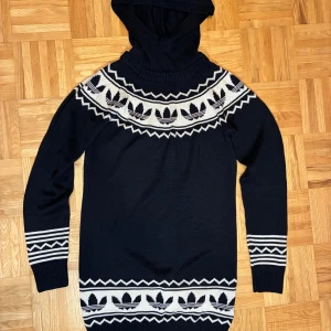 Svart stickad hoodie från Adidas - Svart stickad hoodie från Adidas med vit mönstrad design och ikoniska treklövermönster runt hals och nederkant. Tröjan har långa ärmar, ribbade muddar och en stor huva. Perfekt för dig som gillar streetwear och vill ha något unikt i garderoben.