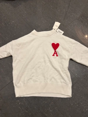 Vit stickad tröja från Ami med hjärta - Vit stickad tröja från Ami med röd hjärtapplikation och bokstaven A på bröstet. Tröjan har ribbad krage, ärmslut och nederkant. Enkel och clean design som ändå sticker ut med den röda detaljen framtill.