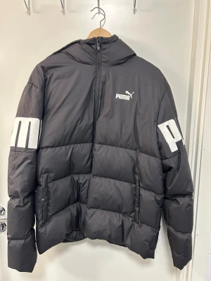 Svart pufferjacka från Puma M - Svart pufferjacka från Puma i storlek M. Jackan har huva, dragkedja framtill och stora vita Puma-loggor på ärmarna. Den är vadderad och har två fickor med dragkedja. Perfekt för kalla dagar och har en sportig vibe.