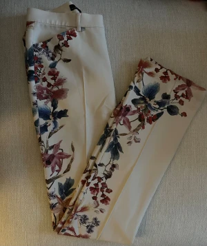 Blommiga vita byxor med raka ben - Snygga vita byxor med färgglatt blommönster längs benen. Byxorna har raka ben och bälteshällor i midjan. Materialet känns mjukt och bekvämt.
