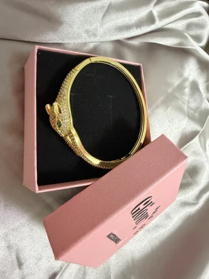 Glittrigt guldarmband med leopardhuvud  - Unikt armband i guldton med leopardhuvud, täckt av glittrande stenar och gröna ögon. Designen är detaljerad och djurinspirerad, perfekt för dig som gillar statement-smycken. Kommer i en rosa presentask från Glow Accessories.