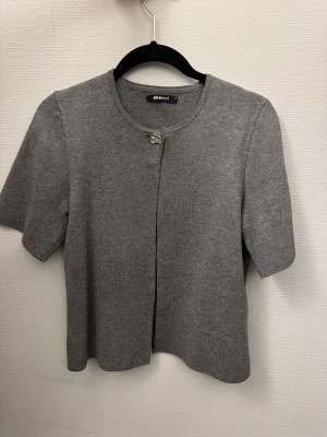 Grå stickad bolerotopp från Gina Tricot - Säljer en stilren grå stickad bolerotopp från Gina Tricot med korta ärmar och rund hals. Toppen har en enkel knäppning med en liten silverfärgad detalj vid halsen. Perfekt för att addera en chill vibe till din outfit.