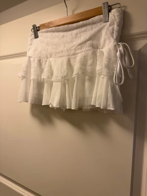Vit volangkjol med spets och knyt S - Supersöt vit kortkjol med volanger och spetsdetaljer. Kjolen har ett lager av mesh och spets upptill, samt ett knytband på sidan för extra stilpoäng. Perfekt för dig som gillar romantiska och trendiga plagg med lite extra fluff.