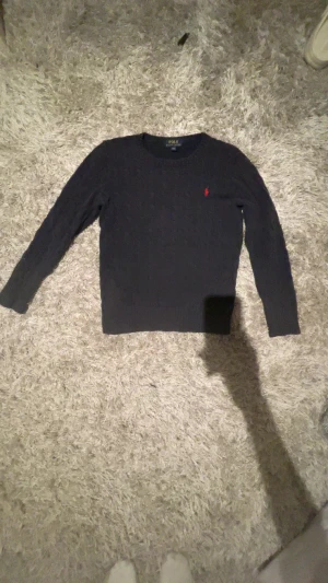 Mörkblå stickad tröja Polo Ralph Lauren - Snygg mörkblå stickad tröja från Polo Ralph Lauren med klassisk kabelstickning och röd broderad logga på bröstet. Rund hals och långa ärmar, perfekt för dig som gillar stilrena och tidlösa plagg. Passar dig som vill ha en normal passform. Passar 160-170cm