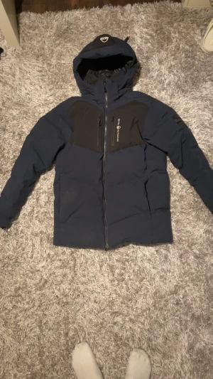 Mörkblå pufferjacka Sail Racing - Mörkblå pufferjacka från Sail Racing med svarta detaljer över axlar och bröst. Jackan har huva med logga, hel dragkedja framtill och två bröstfickor med dragkedja. Perfekt för kalla dagar och har en sportig look.