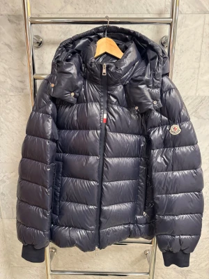 Mörkblå Moncler pufferjacka med huva - Säljer en mörkblå pufferjacka från Moncler med avtagbar huva och ribbade muddar. Modellen heter Cuvellier Giubbotto och liknar Maya men med en fransk flagga vid dragkedjan. Jackan har en blank yta, Moncler-logga på ärmen och dragkedja framtill. Perfekt för kalla dagar och har en riktigt schysst look som sticker ut. Jackan är mycket sparsamt använd. Nypris ligger på 17000