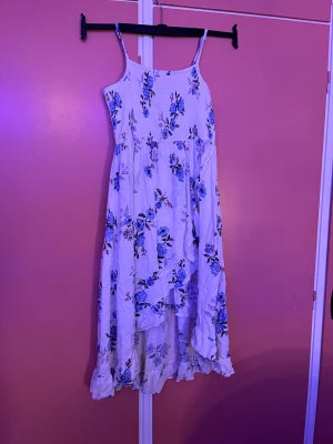 Vit blommig långklänning från Lindex - Superfin långklänning från Lindex i vitt med blått blommönster. Klänningen har smala axelband, smock upptill och volangkant nertill. Perfekt för sommarens varma dagar och har en flowy, luftig känsla. Tillverkad i mjuk bomull.