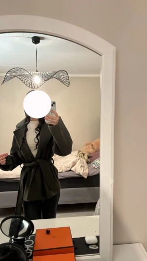 Mörkgrön kappa med bälte - Snygg mörkgrön kappa med breda slag och knytbälte i midjan. Jackan har en loose passform och är perfekt för dig som gillar stilrena och enkla plagg. Materialet känns mjukt och värmande, och den har långa ärmar för extra komfort.