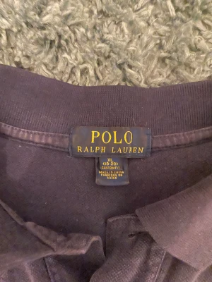 Marinblå pikétröja Ralph Lauren XL - Säljer en marinblå pikétröja från Polo Ralph Lauren i storlek XL (barn storlek). Tröjan har klassisk krage, två knappar och stor vit logga på bröstet samt en vit siffra 3 på ärmen. Materialet är mjuk bomull och passformen är normal. Perfekt för dig som gillar stilren och sportig look.