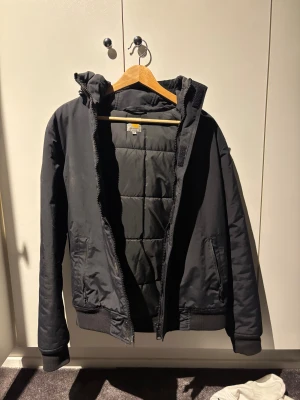 Svart pufferjacka Carhartt M - Svart pufferjacka från Carhartt i storlek M. Jackan har huva, broderad logga på bröstet och quiltad insida. Utrustad med dragkedja och två fickor framtill. Ribbad mudd vid ärmslut och nederkant för extra komfort. Perfekt för kalla dagar. 
