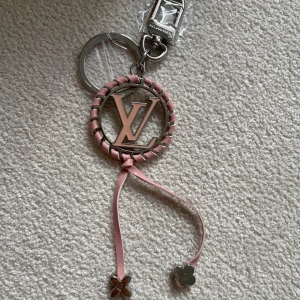  Vuitton rosa och silver nyckelring - Nyckelring från LVuitton med ikoniska-loggan i silver och rosa. Rund form med rosa band flätat runt kanten och två hängande band med blommor i silver. Perfekt som accessoar till väskan eller nycklarna för en lyxig touch.