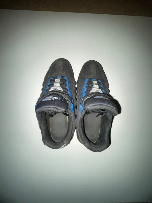 Nike Air Max 95 svart/blå sneakers - Nike Air Max 95 sneakers i svart och blått med grå detaljer. Skorna har synlig Air Max-dämpning, snörning och meshpaneler för extra ventilation. Swoosh-logga på sidan och Air Max-märke på plösen. Snygg vågig design och robust sula för grym komfort.