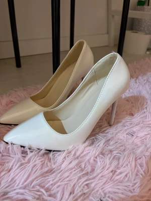 Vita klackar med röd sula, storlek 37 - Säljer ett par stilrena vita klackar med glansig finish och röd undersida. Skorna har spetsig tå och hög klack som ger en snygg siluett. insidan är ljusbeige, skon har en liten fläck. Perfekt för dig som gillar klassisk och elegant stil.