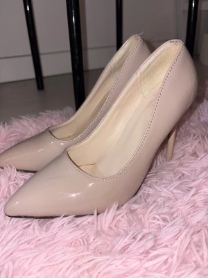 Beige klackar med spetsig tå, strl 37 - Snygga beige klackar med spetsig tå och hög klack. Skorna har en glansig finish. Insidan är ljus och sulan har en röd detalj undertill. Perfekt för dig som gillar stilrena och eleganta skor.