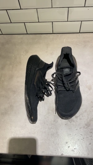 Svarta Adidas Ultraboost sneakers - Svarta Adidas Ultraboost sneakers med stickad ovandel och platt sula. Skorna har klassisk snörning och de ikoniska tre ränderna på sidan i plast. Sulan är grov och ger bra dämpning, perfekt för dig som gillar sportig stil och komfort.