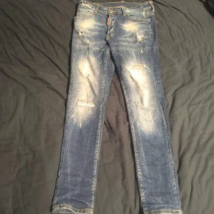 Dsquared2 slitna blå jeans strl 42 - Säljer ett par Dsquared2 jeans i blå tvätt med slitningar och distressed detaljer. Modellen har klassisk femficksdesign, raka ben och snygga kontrastsömmar. Röd Dsquared2 Denim-patch baktill. äkta dsq jeans vart med i g1oactore music video (pierroo)