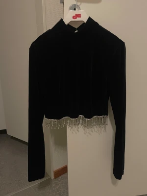 Svart topp - Säljer en svart croppad topp i mjuk sammet med lång ärm och hög hals. Nederkanten är dekorerad med glittrande pärlor. Använd endast en gång.