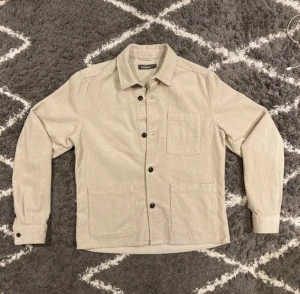 Beige overshirt från Dressmann - Snygg beige overshirt från Dressmann i mjuk manchester. Jackan har klassisk krage, tre stora fickor framtill och svarta knappar. Perfekt att slänga på över en t-shirt för en chill och stilren look. Jag är 180 och väger 68kg och den sitter bra.
