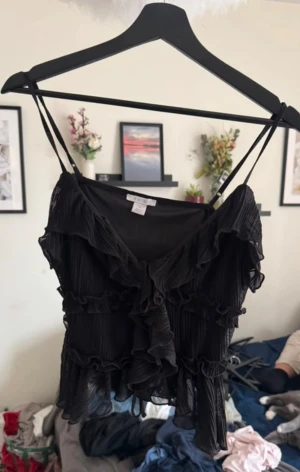 Svart volangig linnetopp från Amisu - Säljer en svart linnetopp från Amisu med tunna axelband och flera lager volanger. Toppen är plisserad och har en luftig, flowy känsla som ger en cool och feminin vibe. Perfekt för dig som gillar detaljer och vill sticka ut lite extra.