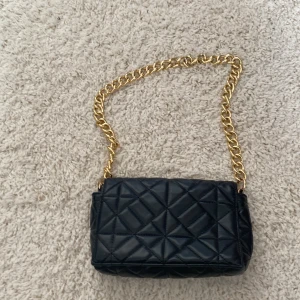Svart quiltad axelväska med kedja - Snygg svart axelväska med quiltat mönster och grov guldfärgad kedja som axelrem. Väskan är rektangulär och har ett innerfoder i vinrött. 