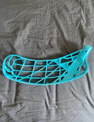 Oxdog innebandyblad - Bilderna visar ett innebandyblad i turkos färg med förstärkning i kolfiber. Bladet har ett modernt, öppet designmönster och ser ut att vara i gott skick med endast mindre ytliga spår av användning. Perfekt för dig som vill ha ett lätt och stabilt blad till din innebandyklubba.