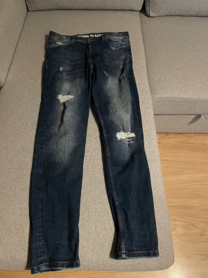Mörkblå Hoodrich jeans med slitningar - Säljer ett par mörkblå jeans från Hoodrich med coola slitningar och distressed detaljer på benen. Jeansen har broderad logga på bakfickan och texten 'FROM NOTHING TO SOMETHING' på insidan av midjan. Klassisk femficksmodell med raka ben och snygg tvätt.