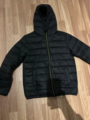 Svart pufferjacka med huva - Svart pufferjacka med huva och horisontella sömmar. Jackan har dragkedja framtill i kontrasterande färg och två stora fickor. Perfekt för kyliga dagar och har en stilren, enkel look.