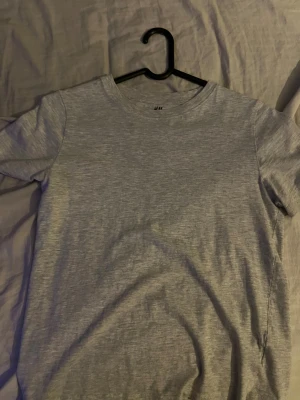 Grå basic t-shirt från H&M - En enkel och stilren grå t-shirt från H&M Basic, tillverkad i mjuk ekologisk bomull. Modellen har rund hals och korta ärmar, perfekt för en chill och clean look. Passar dig som gillar minimalistisk stil och bekväma plagg.