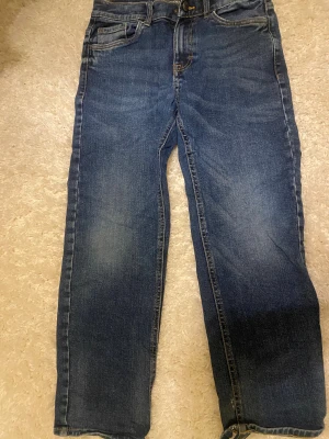 Blå jeansbyxor från Lindex, strl 140 - Snygga blå jeansbyxor från Lindex i klassisk femficksmodell. Byxorna har rak passform och normal midja, med slitningar och tvättade detaljer för en cool look. Materialet är jeans med kontrastsömmar och knappstängning framtill.