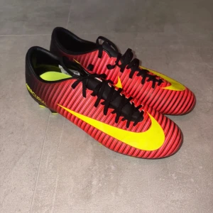 Nike Mercurial Fotbollsskor - Säljer ett par Nike Mercurial fotbollsskor i storlek 40.5. Skorna har en snygg randig design i rött och svart med gula detaljer och snörning. Yttersulan har gula dobbar för gräsplan. Perfekta för dig som vill sticka ut på planen. 