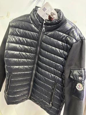 Mörkgrå pufferjacka från Moncler - Snygg mörkgrå pufferjacka från Moncler med quiltad front och stickade ärmar. Jackan har dragkedja framtill, två sidofickor med dragkedja och en Moncler-logga på ärmen. Perfekt för dig som gillar exklusiv streetstyle.