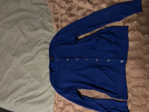 Kornblå kofta från Brandtex Classic - Kofta i kornblå färg från Brandtex Classic, storlek M. Den har långa ärmar, rund hals och knappar hela vägen framtill. Två fickor framtill och ribbade muddar vid ärmslut och nederkant. Perfekt för dig som gillar klassisk stil med en färgklick.