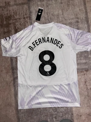 Manchester United B. Fernandes tröja M - Snygg Manchester United fotbollströja från Adidas med B. Fernandes och nummer 8 på ryggen. Tröjan är vit med ljuslila grafiskt mönster, svarta detaljer och klassiska Adidas-ränder på axlarna. V-ringad krage och tillverkad i lätt polyester med AEROREADY-teknologi.