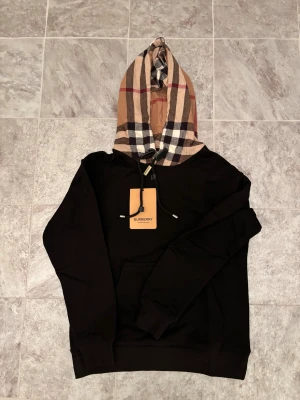 Svart Burberry hoodie med rutig huva - Svart hoodie från Burberry med klassisk rutig huva i beige, svart, vitt och rött. Tröjan har snörning vid huvan och en stor magficka framtill. Perfekt för dig som vill ha en stilren men ändå ikonisk look.