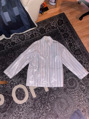Overshirt acne studios - Snygg randig skjorta från Acne Studios i vitt och ljusblått med dragkedja framtill. Skjortan har klassisk krage och långa ärmar. Materialet är 100% bomull med en blank polyuretan-coating som ger en unik glansig finish. Perfekt för dig som gillar statement-plagg.