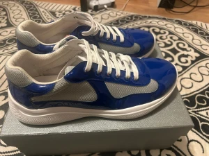 Blå och grå sneakers från Prada - Säljer ett par snygga Prada sneakers i blått och grått med vita snören och detaljer i rött. Ovandelen är i mesh och lackad syntet, med rund tå och platt sula. Ikonisk Prada-logga på plösen och sula. Perfekt för dig som gillar exklusiva märken och sportig stil.