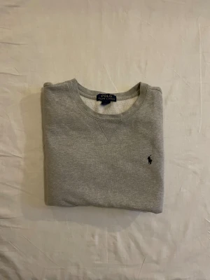 Grå sweatshirt från Polo Ralph Lauren - Snygg grå sweatshirt från Polo Ralph Lauren med klassisk rund hals och broderad logga på bröstet. Tröjan har långärmade muddar och är gjord i mjuk bomullsmix som är skön att bära. Perfekt för chill dagar och enkel att matcha med jeans eller joggers.