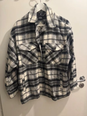 Rutig overshirt från Zara i grått - Säljer en sjukt snygg rutig overshirt från Zara i grå, svart och vit. Jackan har två stora bröstfickor med knappar, klassisk krage och en loose passform. Materialet är mjukt och lite fluffigt, perfekt för lager-på-lager. Passar dig som gillar street och chill stil.