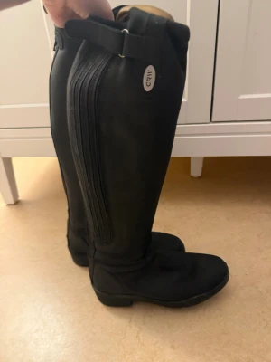 Svarta ridstövlar från CRW i skinn - Säljer ett par svarta ridstövlar från CRW i skinn. De har dragkedja baktill, justerbart spänne upptill och elastisk panel längs skaftet för extra komfort. Klassisk design med låg klack och rund tå, perfekta för ridning. Pris kan diskuteras!