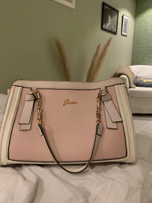Guess ljusrosa handväska med kedja - Snygg handväska från Guess i ljusrosa och beige med guldfärgade kedjedetaljer och logga framtill. Väskan har flera fack, dragkedja och innerfoder med G-mönster. Väskan är lite sliten på insidan samt banden på insidan har tappat lite färg. 