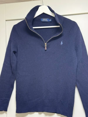 Marinblå half zip tröja Polo Ralph Lauren - Äkta Polo Ralph Lauren halvzip-tröja i mörkblått stickat material. Tröjan har en hög krage med metallblixtlås och det klassiska ljusblå broderade Polo-logot på bröstet. Modellen är något mindre i storlekenså den passar som en smäck i storleken xs. Tänk på det vid köp. Den är i fint skick och det är bara att höra av sig vid minsta fundering eller fråga.