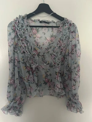 Blus Zara - Supersöt blommig blus från Zara i ljusblått med volangdetaljer längs v-ringningen och ärmsluten. Tunt och transparent tyg med pastellfärgade blommor, passar S/M