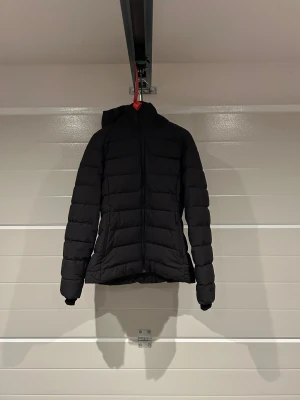 Svart pufferjacka från Everest - Svart pufferjacka från Everest med huva och dragkedja framtill. Jackan har en quiltad look med horisontella sömmar och två fickor med dragkedja. Perfekt för kalla dagar när du vill hålla värmen och ändå se stilren ut.