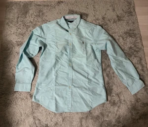 Ljusblå skjorta från Ralph Lauren - Snygg ljusblå skjorta från Ralph Lauren med klassisk button-down krage och broderad logga på bröstet. Skjortan har långa ärmar, vita knappar och en stilren passform som ger en fräsch look. Perfekt för dig som gillar stilrena plagg med premiumkänsla.