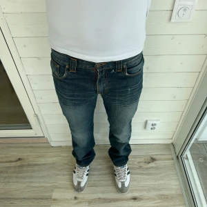 Blå jeans från Nudie Jeans - Säljer ett par klassiska blå jeans från Nudie Jeans med orangea sömmar och läderpatch bak i midjan. Jeansen har en slitning bredvid gylfen. Modellen är 183cm. Skriv vid funderingar💬
