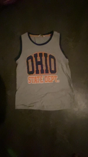 Grå linnetopp med OHIO-tryck från M&S - Säljer en grå linnetopp från M&S med stort OHIO STATE DEPT.-tryck i blått och orange på bröstet. Toppen har mörkblå kant runt hals och ärmhål, och är tillverkad i mjuk bomull. Perfekt för dig som gillar sportig och avslappnad stil.