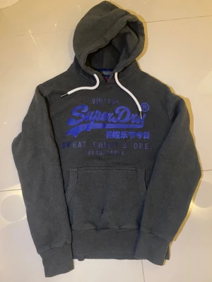 Mörkgrå Superdry hoodie med blått tryck - Säljer en mörkgrå hoodie från Superdry med stort blått tryck på bröstet och vit snörning i huvan. Tröjan är vintage och väldigt svårt att få tag på, passar som ett M