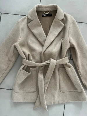 Beige kappa med bälte från Zara XS - Stilren beige kappa från Zara i storlek XS. Jackan har klassisk krage, knytskärp i midjan och stora fickor framtill. Materialet är mjukt och känns lyxigt, perfekt för dig som gillar en clean och snygg look. Lite lätt kopprig men ingenting man lägger märke till, går att få bort enkelt:)