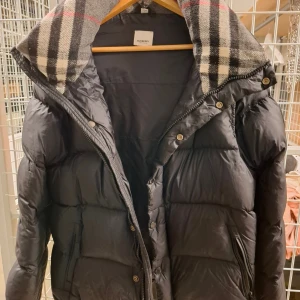 Svart pufferjacka från Burberry S - Svart pufferjacka från Burberry med klassiskt rutigt foder i huvan. Jackan har dragkedja och knappar framtill, samt Burberry-logga på ärmen. Perfekt för kalla dagar med sin vadderade design och snygga detaljer.