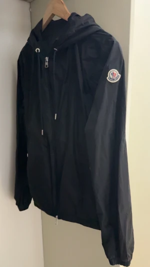 Svart Moncler vindjacka med huva - Svart vindjacka från Moncler med huva och snygga vita och röda ränder på insidan av huvan. Jackan har dragkedja framtill, snörning vid huvan och Moncler-logga på ärmen. Materialet är lätt och glansigt, perfekt för blåsiga dagar. Inte använt den mycket alls köpte den i september 